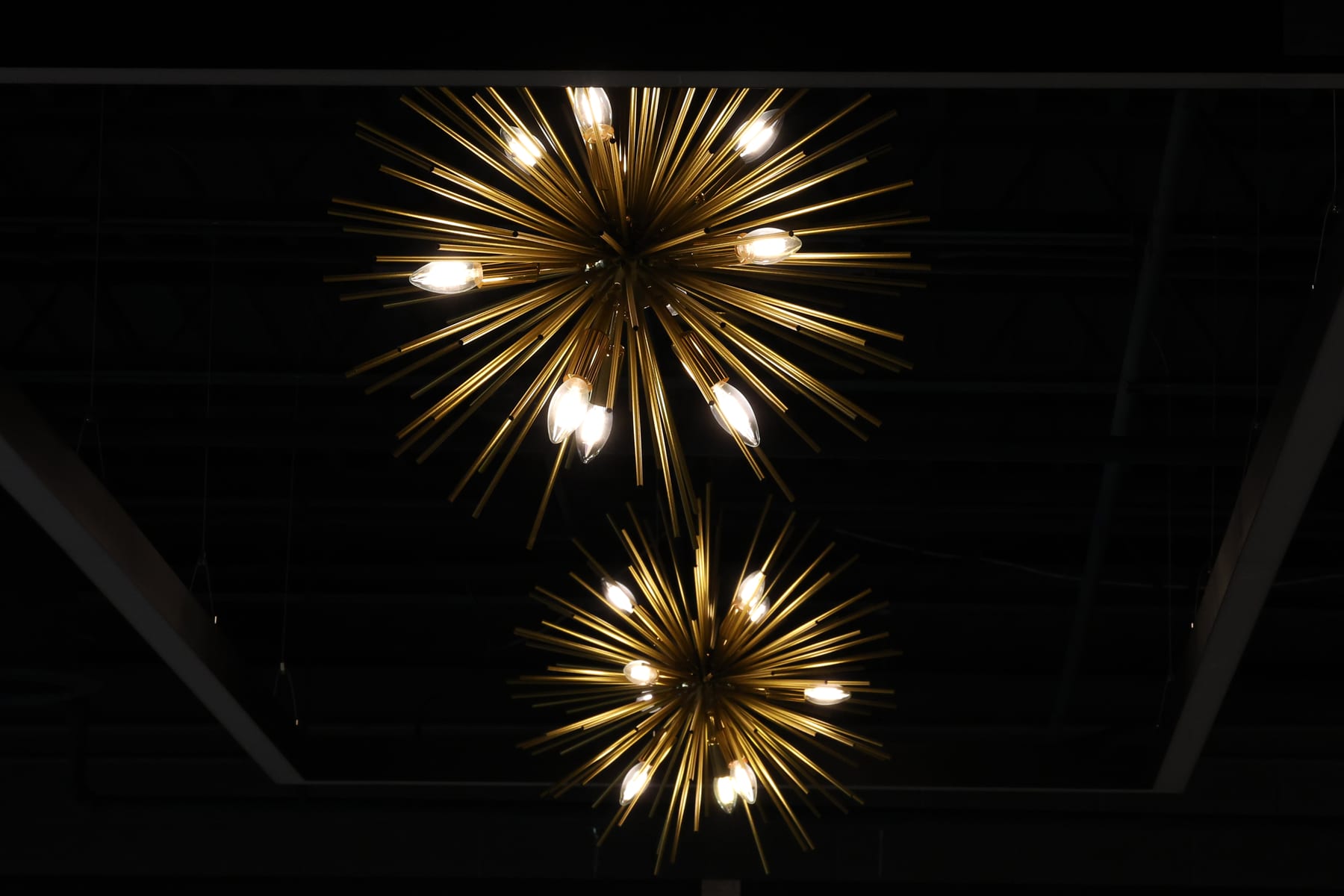 A pair of brass starburst chandeliers, lit, against the dark ceiling